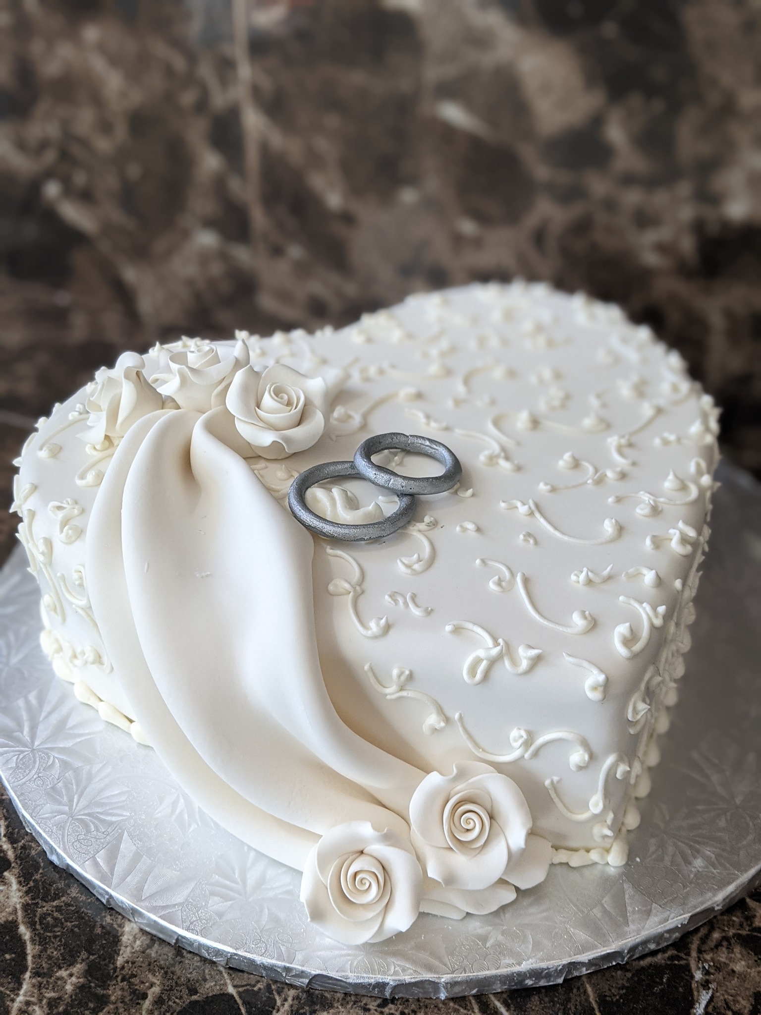 Heart-shaped white wedding cake with elegant scrollwork, sugar roses, and silver ring topper