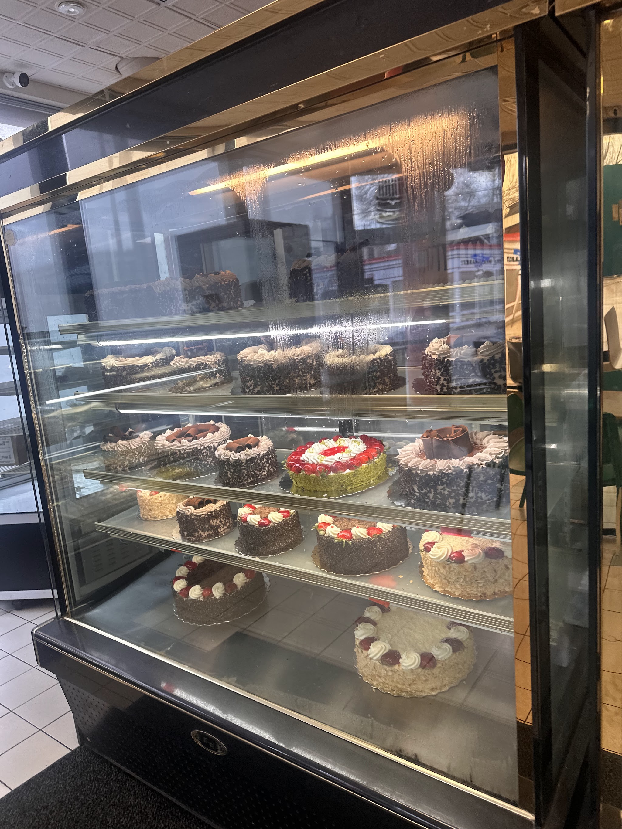 Glass display case showcasing four shelves of European-style cakes including Black Forest, strawberry torte, and cream cakes