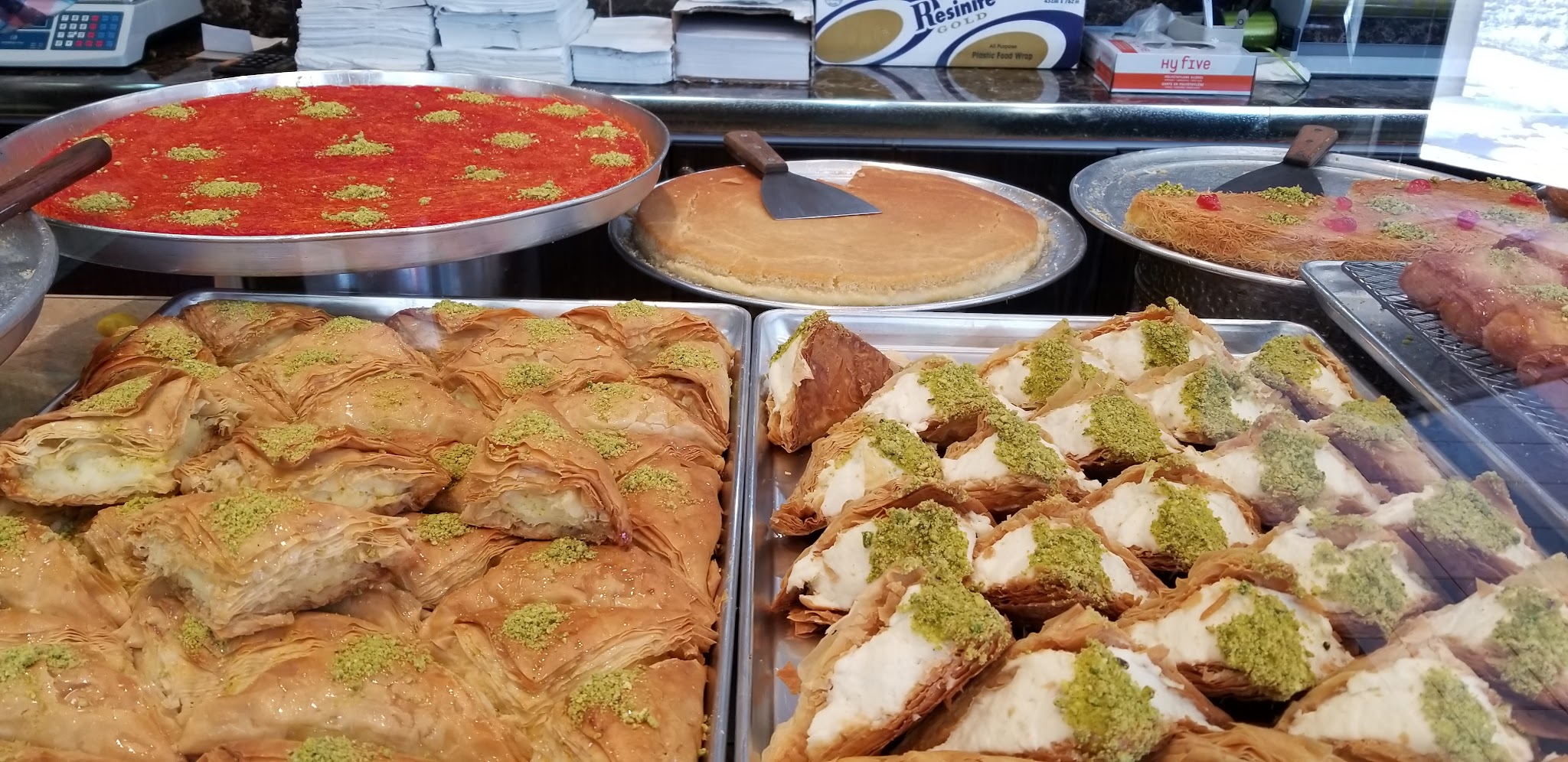Golden baklava rolls topped with crushed pistachio alongside kunafa and assorted Middle Eastern pastries
